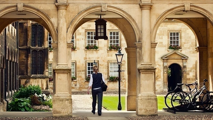 A young woman walks through an archway in a historic Cambridge college courtyard.