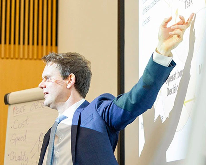 A professional speaking while gesturing towards presentation slides in a classroom setting.