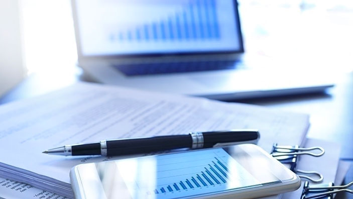 A smartphone displaying a graph, a pen and documents on a table with a laptop in the background.
