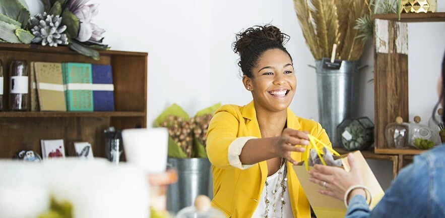 A smiling saleswoman in a yellow blazer hands over a shopping bag to a customer. Behind her are flowers in large metal vases, and notepads and pretty glass jars on a couple of makeshift shelves.
