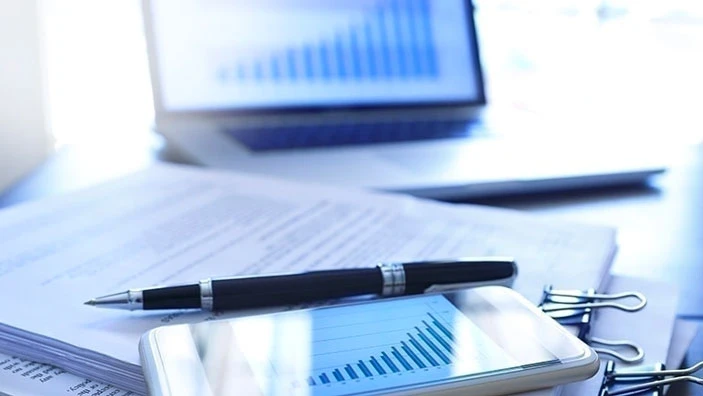 A smartphone displaying a graph, a pen and documents on a table with a laptop in the background.