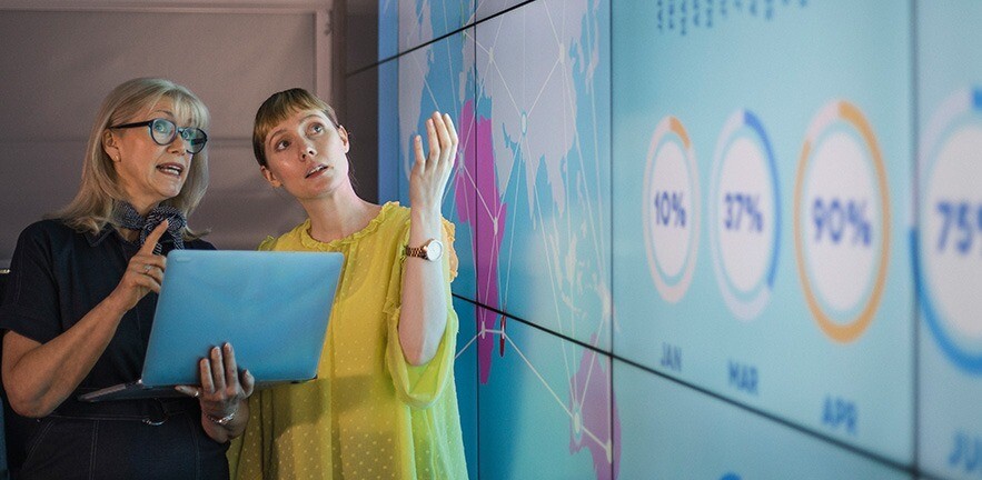 An experienced woman mentors a female colleague, the mature woman is holding a laptop as they debate data from an interactive display; they are both wearing smart casual clothing.