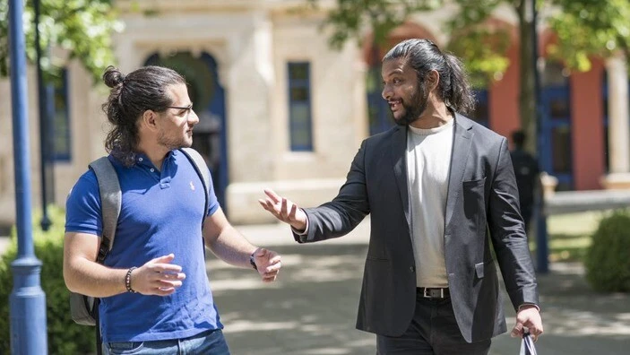 Two men engage in conversation while walking on a sunny campus path, surrounded by greenery and Business School buildings.