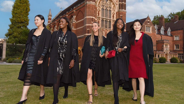 MSt in Entrepreneurship graduates in academic gowns smile and walk together on campus grounds.