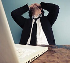Stressed man sitting at a desk with a laptop, head in hands.