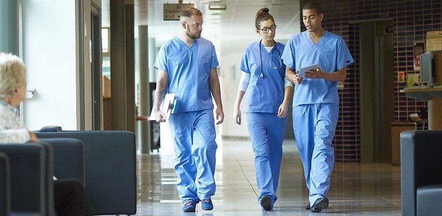 Three junior doctors walking along a hospital corridor discussing case and wearing scrubs.
