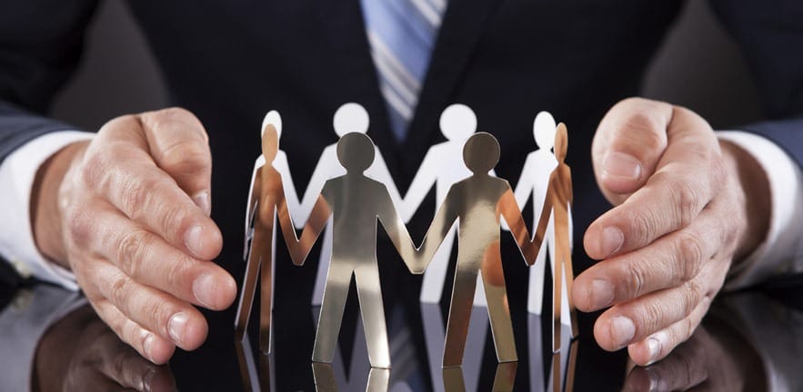 Businessman's Hands Protecting Team Of Paper People On Desk