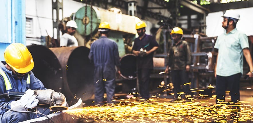 Metal grinder patiently practicing his craft on a large metal pipe. With a group of factory employees working in the background. Trying to lift a large metal pipe with a factory crane.