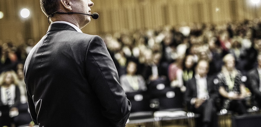Entrepreneur giving a speech in a lecture hall. Audience visible in the background.