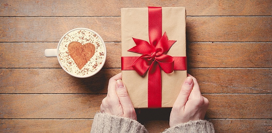 Female hands holding gift box and cup of coffee on wooden table. High angle point of view