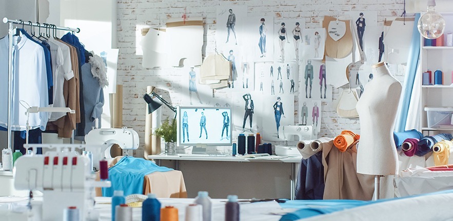 Shot of a Sunny Fashion Design Studio. We See Working Personal Computer, Hanging Clothes, Sewing Machine and Various Sewing Related Items on the Table, Mannequins Standing, Colorful Fabrics.