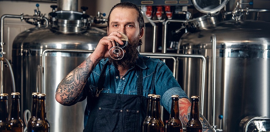 Portrait of tattooed, bearded hipster male manufacturer tasting beer in the microbrewery.