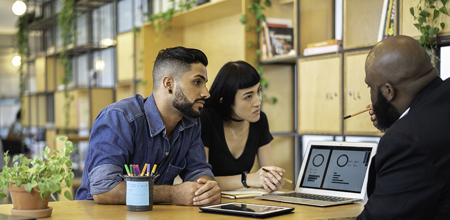 Person discussing an online platform with two other people, a laptop is in shot on the desk.