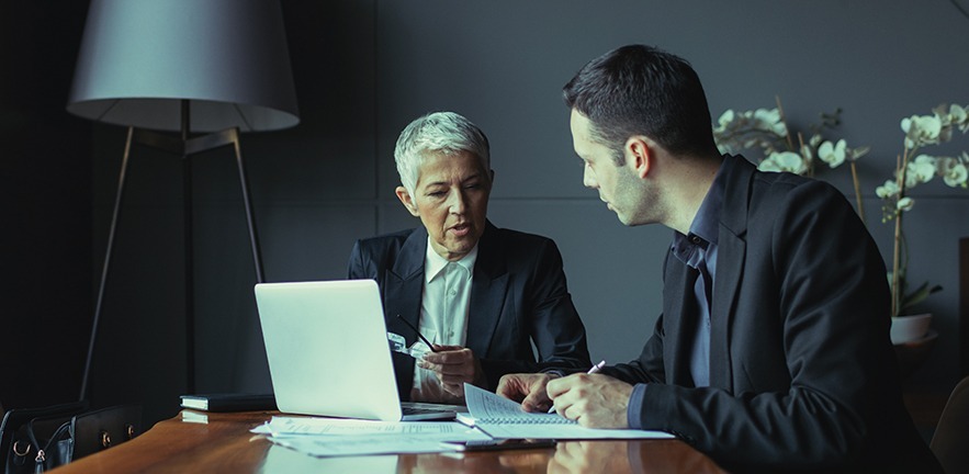 A businesswoman at a laptop discusses something with her assistant.