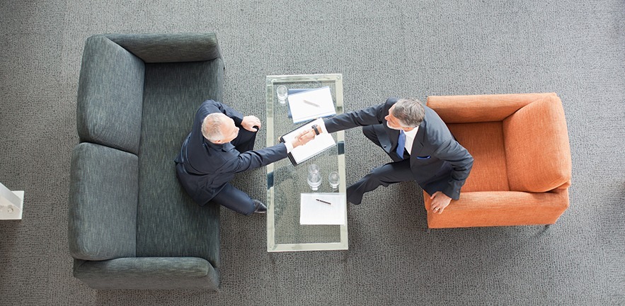 Businessmen shaking hands across coffee table in lobby.