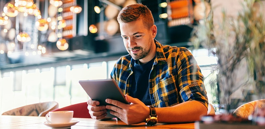 Smiling man using tablet for work. In front of him on a table cup of coffee, in a cafe setting.