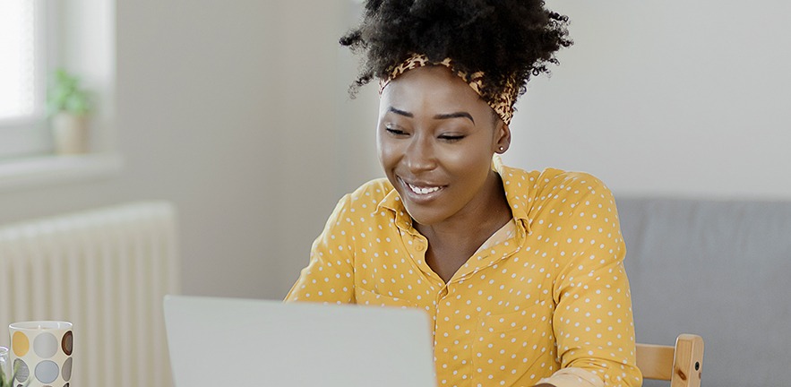 A smiling businesswoman in a yellow shirt smiles at her laptop.