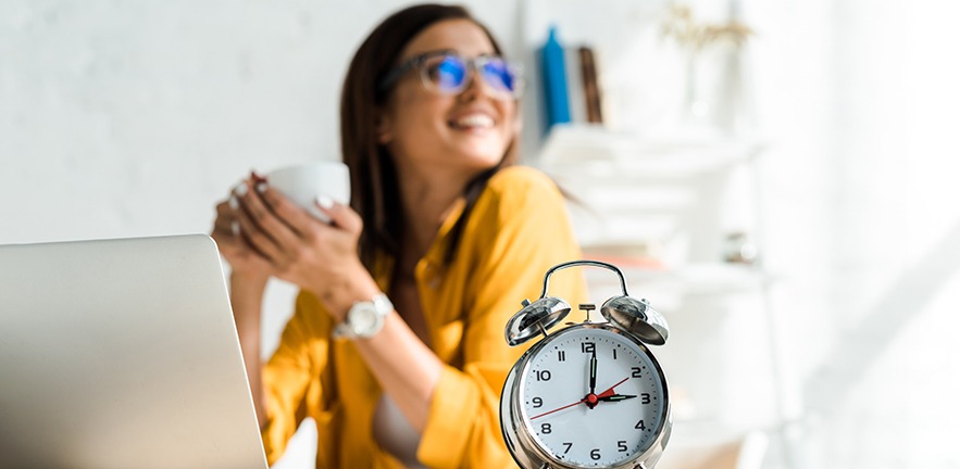 Selective focus of an alarm clock and happy worker having coffee break in home office with laptop.