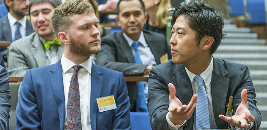 Two MBA classmates sitting in a lecture theatre during and intent discussion as others watch.