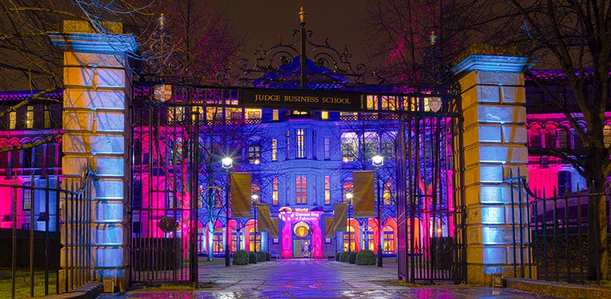 Front facade of Cambridge Judge Business School lit up with colourful lights in support of Cancer.