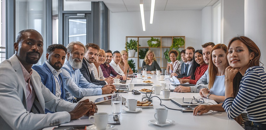 Comprehensive viewpoint of diverse group of business executives sitting together at conference table and looking at camera.