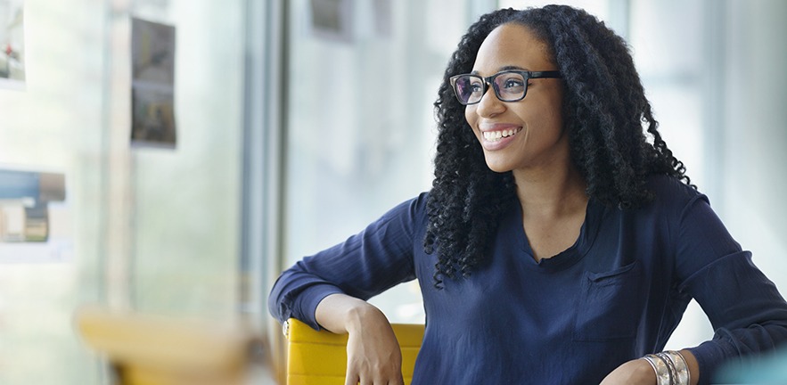 Portrait of young woman entrepreneur smiling and looking happy while sitting on a brightly coloured chair in a naturally lit contemporary office surrounded by glass windows.