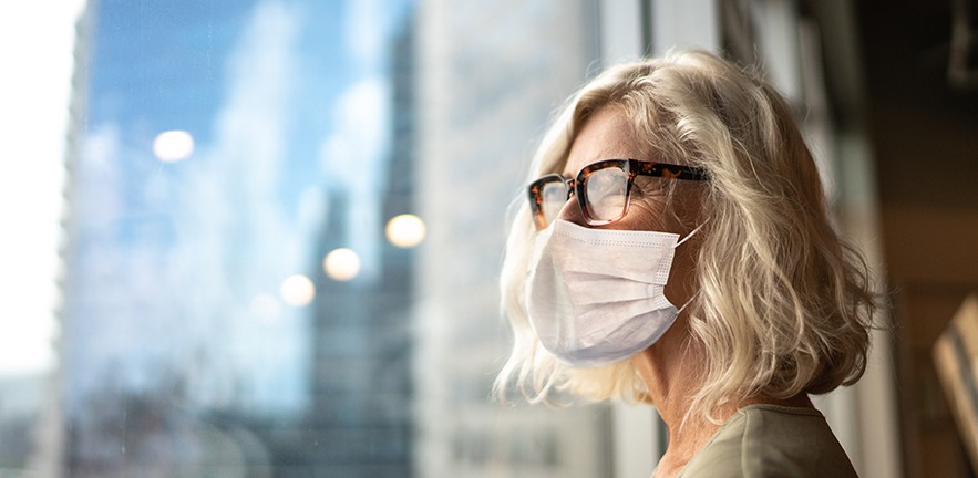 Businesswoman wearing a face mask as she stares into the distance out of an office block window.