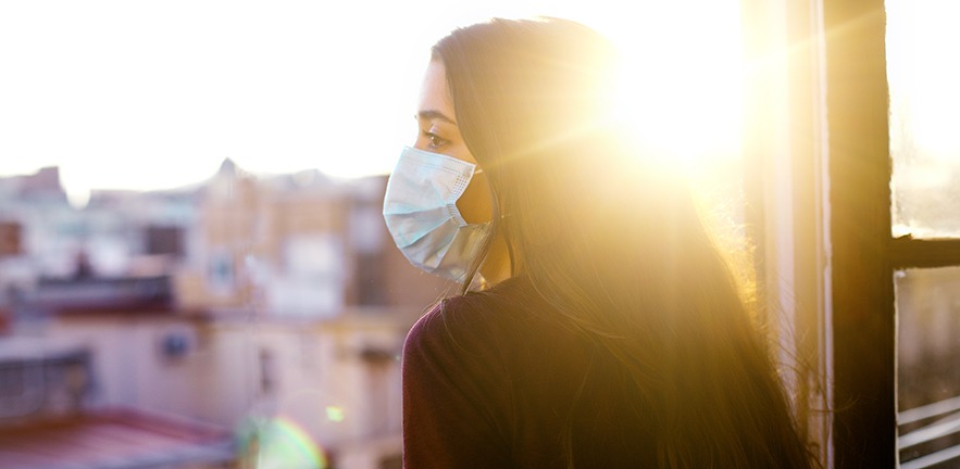 Woman wearing protective mask, looking at the city from the window during coronavirus qurantine in Barcelona. Nice sunset with the sun shining in the sky.