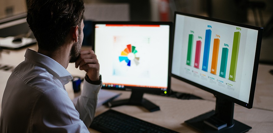 Man doing market research, looking at at data and graphs on two computer screens.