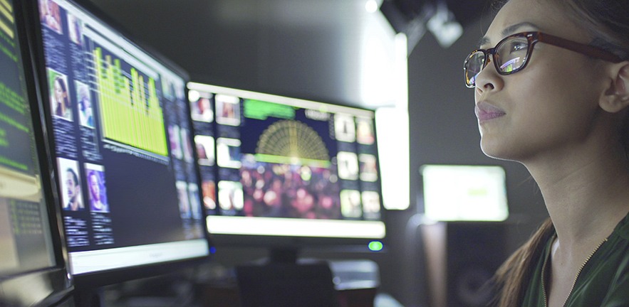 Close up of a young Asian woman sitting down at her desk looking at three large computer monitors displaying data.