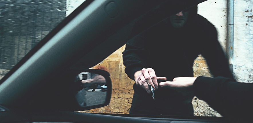 A man in a car sticks his hand out of the window to exchange cash in return for street drugs from a man in black, his face obscured.