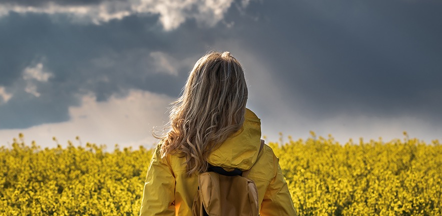 A female hiker in a bright yellow waterproof jacket stands in a rapeseed field and looks at the thunderstorm approaching.