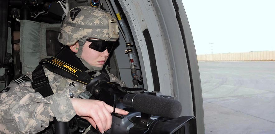 Women in military gear with video camera looking out of helicopter.