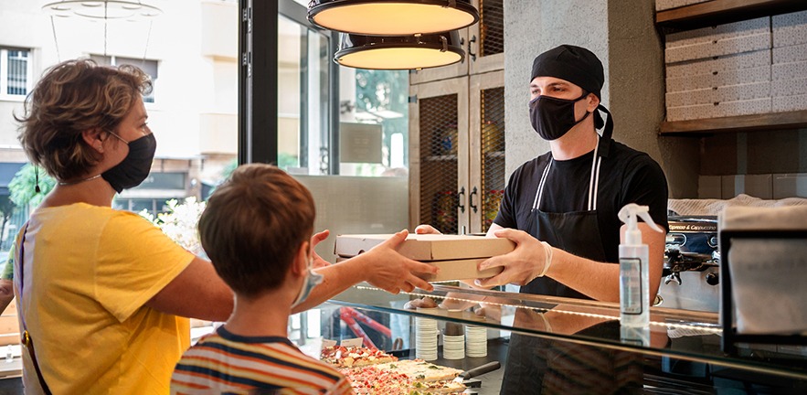 Male checkout server wearing protective gloves and handing fresh pizza in pizza boxes, boy is holding paper bag, both customers is wearing protective masks during Covid-19 pandemic era.