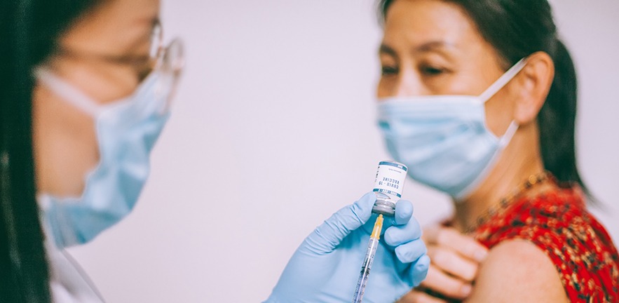 Medical staff member preparing a COVID-19 vaccine with a syringe injection while patient sits patiently with her bare arm out.