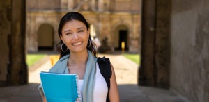 Portrait of a female student at the front of one of the University's Colleges holding a notebook and looking at the camera smiling