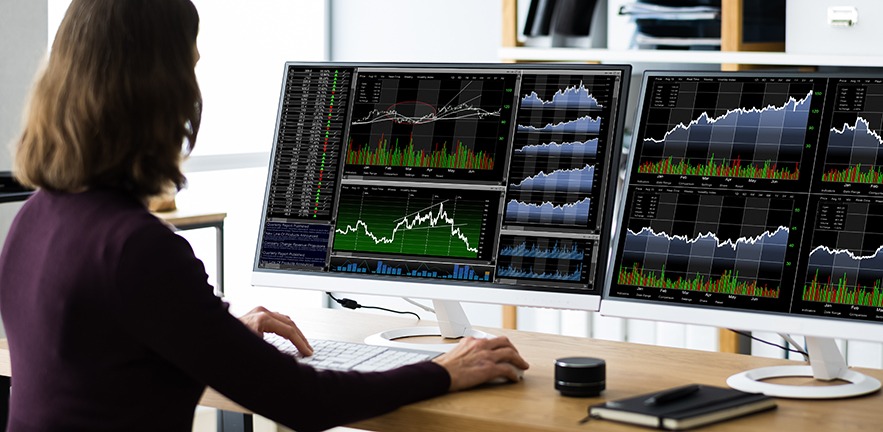 Financial trader facing two computer monitors as she analyses the data and graphs in front of her.