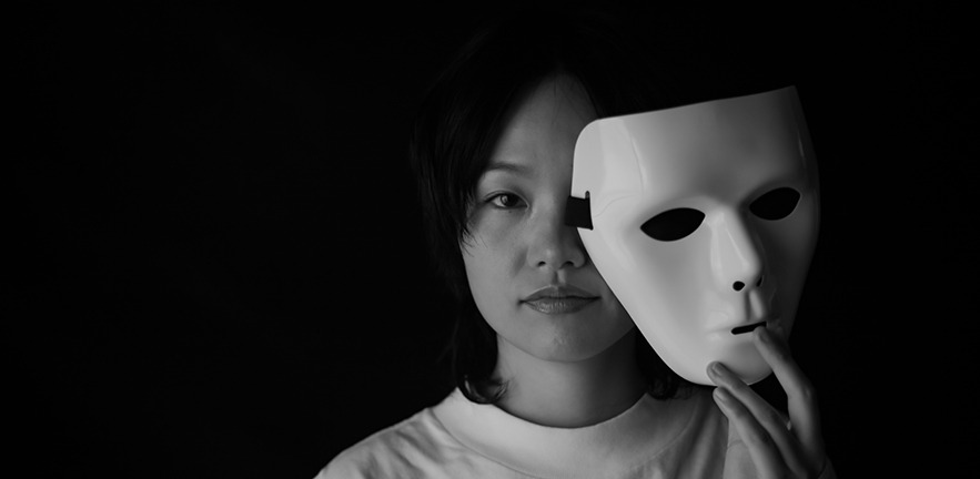 Black and white photo of a woman holding a mask away from her face to reveal a serious expression.