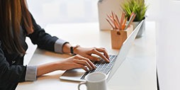 Cropped image of creative woman hands typing on a computer laptop that putting on a white working desk surrounded by a coffee cup, pencil holder, potted plant, and file folder.