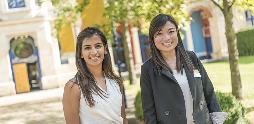 Two MBA candidates smile directly at the Camera while in front of Cambridge Judge Business School on a sunny day.