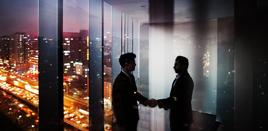 Businessmen shaking hands in a darkened office at night with city lights visible through the window.