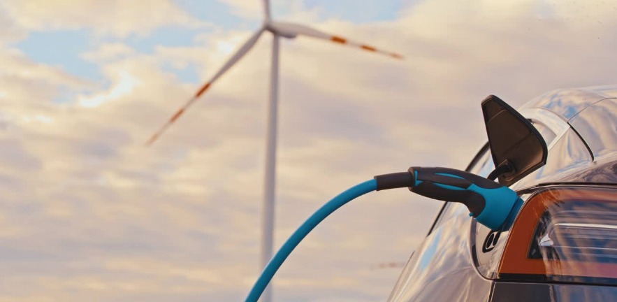 Rear corner of an electric car with charging cable attached. A wind turbine can be seen in the distance.