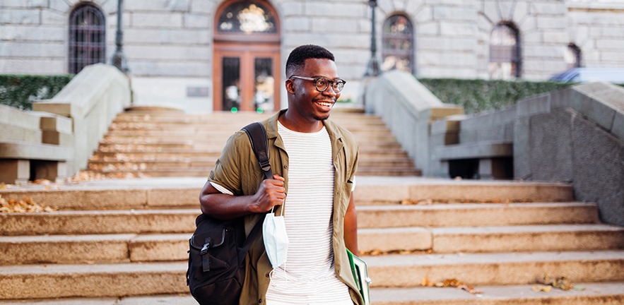 Student smiling and carrying books and a backpack as he walks down his college steps.