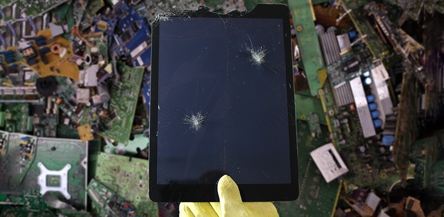 A gloved hand holds up a tablet with smashed screen, broken and deconstructed electronic items lay scattered in the background.