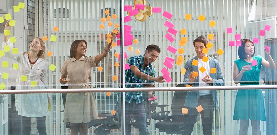 View through a window into an office, where entertained office workers are sticking colourful adhesive notes on the glass.