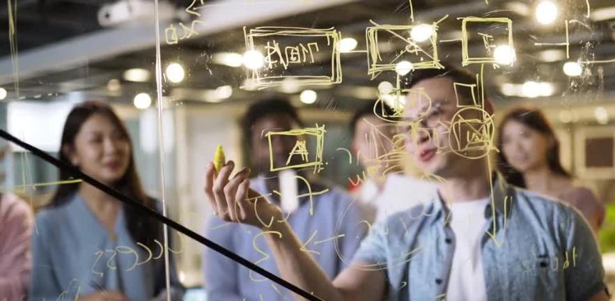 A man in a shortsleeved shirt and glasses uses a yellow crayon to draw a diagram on a clear perspex board to illustrate something to his colleagues.