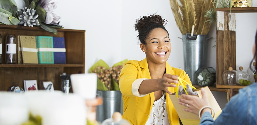 A smiling saleswoman in a yellow blazer hands over a shopping bag to a customer. Behind her are flowers in large metal vases, and notepads and pretty glass jars on a couple of makeshift shelves.