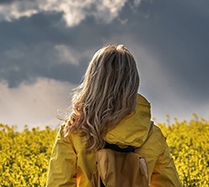 A female hiker in a bright yellow waterproof jacket stands in a rapeseed field and looks at the thunderstorm approaching.