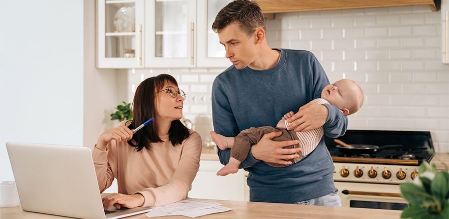 Young mother using laptop in kitchen, young father holds their baby as they discuss plans.