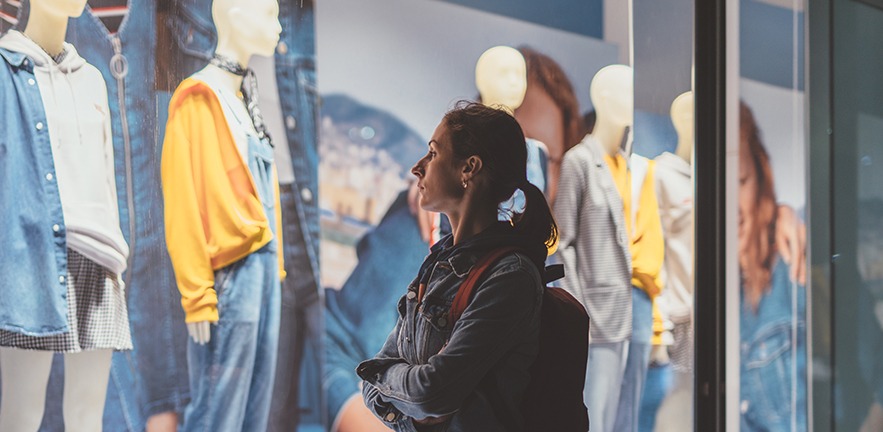 Girl stands outside a clothes shop and looks at the outfits through the window.